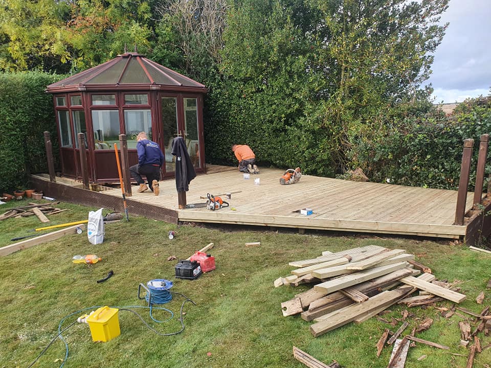 Decking installation in progress with workers, tools, and materials, showcasing a newly built wooden deck adjacent to a conservatory in a garden setting.