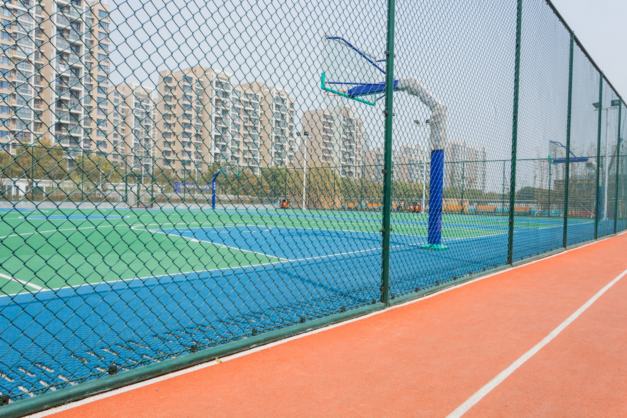 Multi-use games area (MUGA) fencing surrounding a sports court with basketball hoop, featuring vibrant blue and green surfaces, and residential buildings in the background.