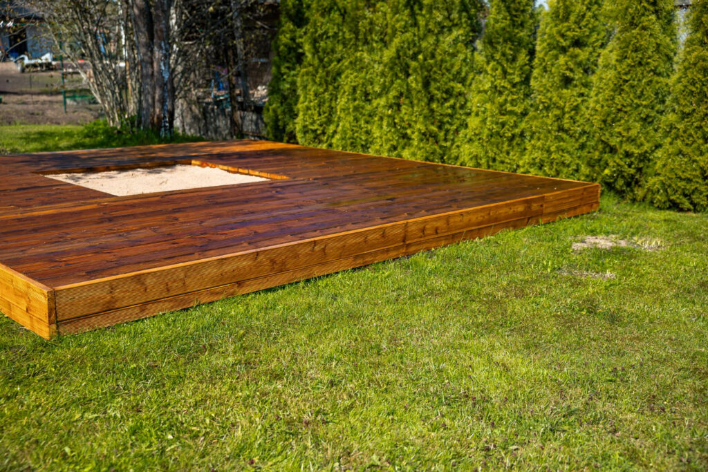 Wooden decking platform with sand pit area, surrounded by green grass and conifer trees, showcasing CK Fencing Contractors' professional decking installation for outdoor spaces.
