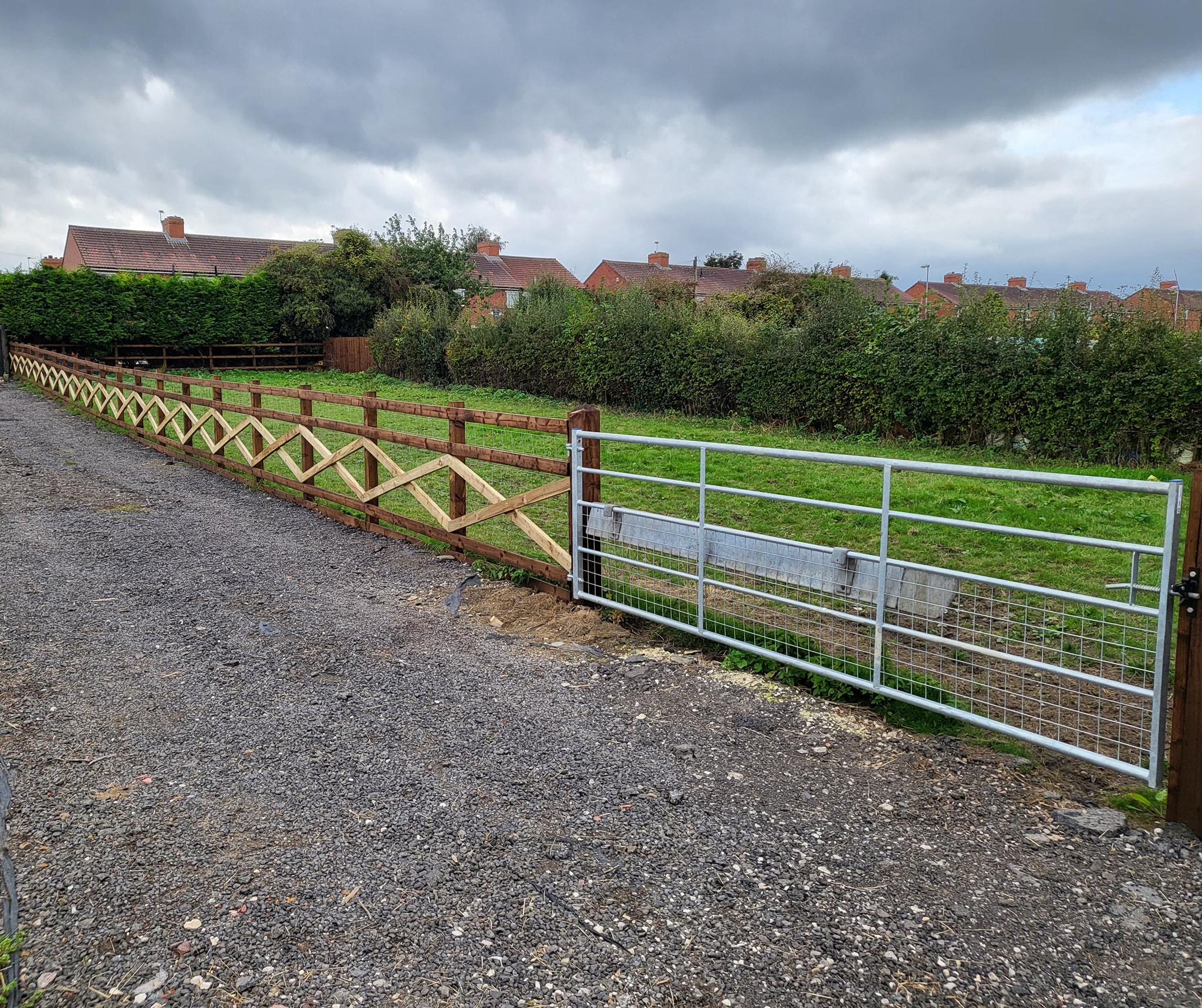 Post and rail fencing along a gravel driveway, featuring a metal gate, surrounding greenery, and residential houses in the background, illustrating agricultural fencing solutions for rural properties.