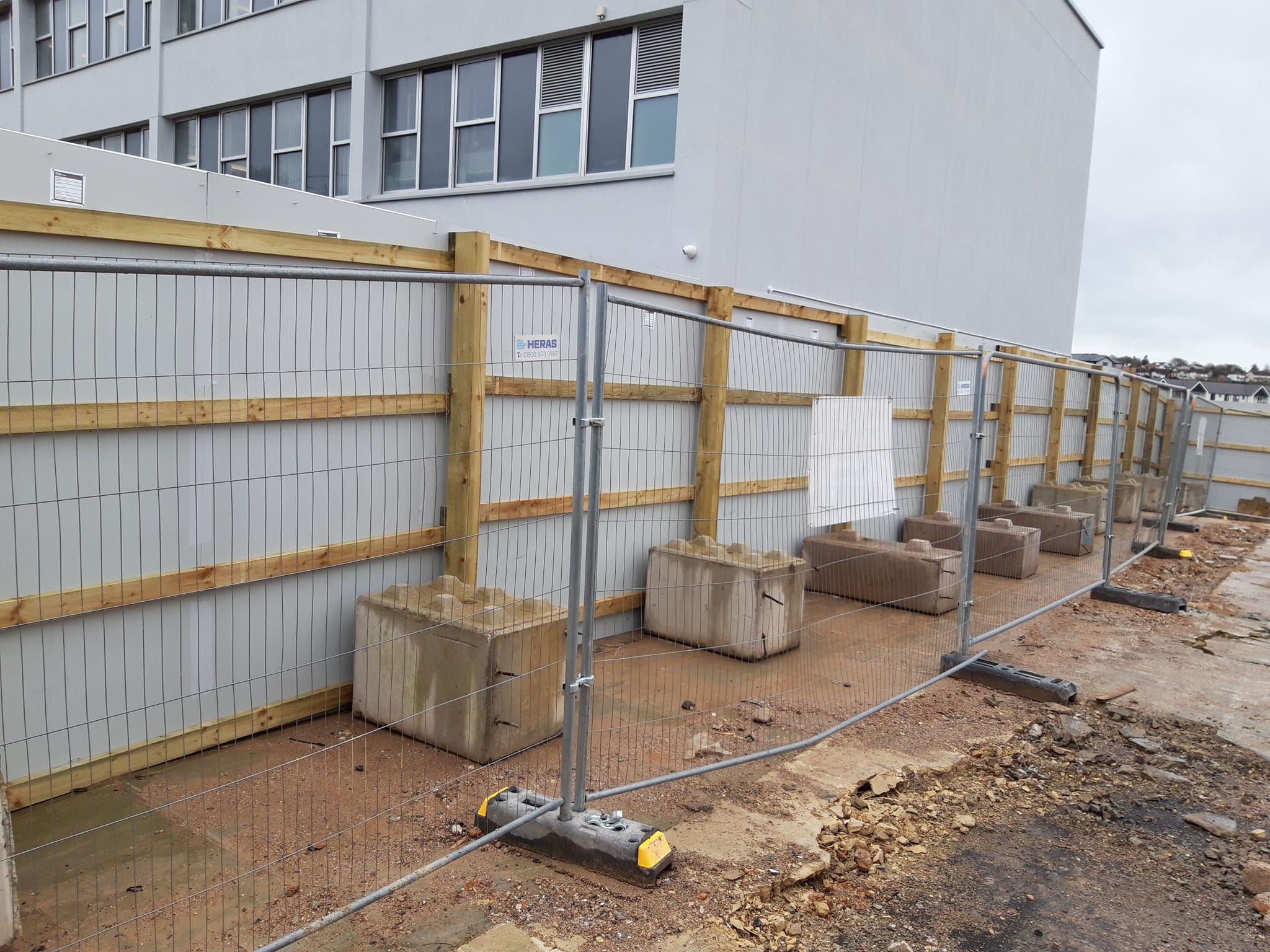 Construction site hoarding with wooden and metal fencing, concrete blocks for stability, and a building in the background, illustrating site security and compliance with UK safety standards.
