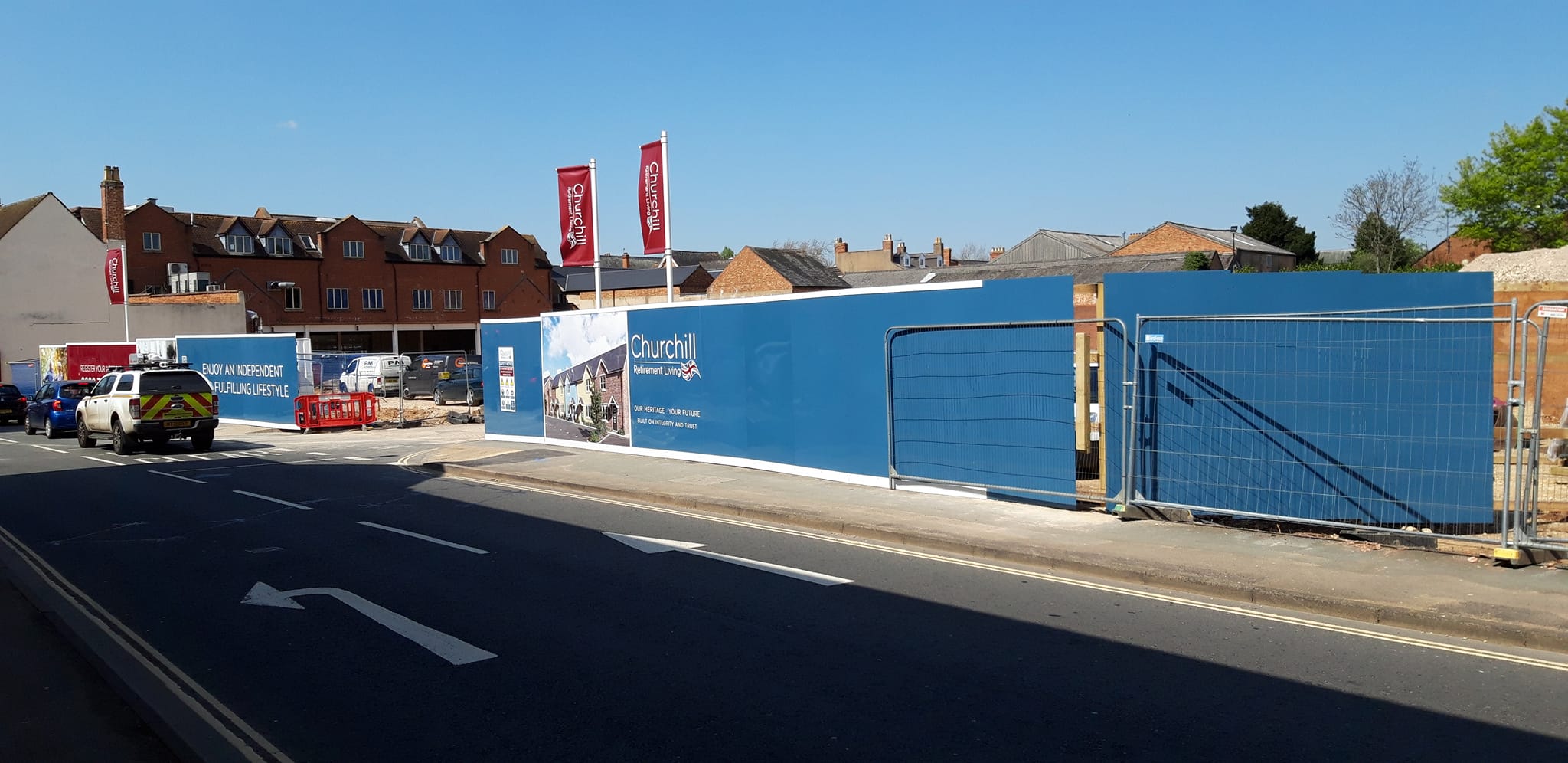 Construction site hoarding featuring blue panels and signage for Churchill Retirement Living, alongside a vehicle and construction equipment, set against a clear blue sky.