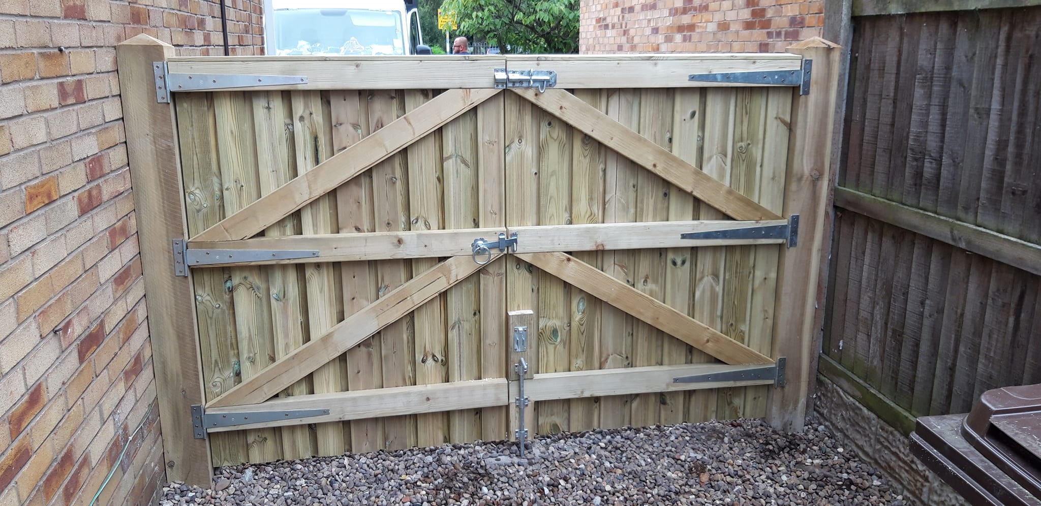 Wooden garden gate with diagonal bracing and metal fittings, set against a brick wall and gravel pathway, showcasing CK Fencing Contractors' craftsmanship in gate installation.