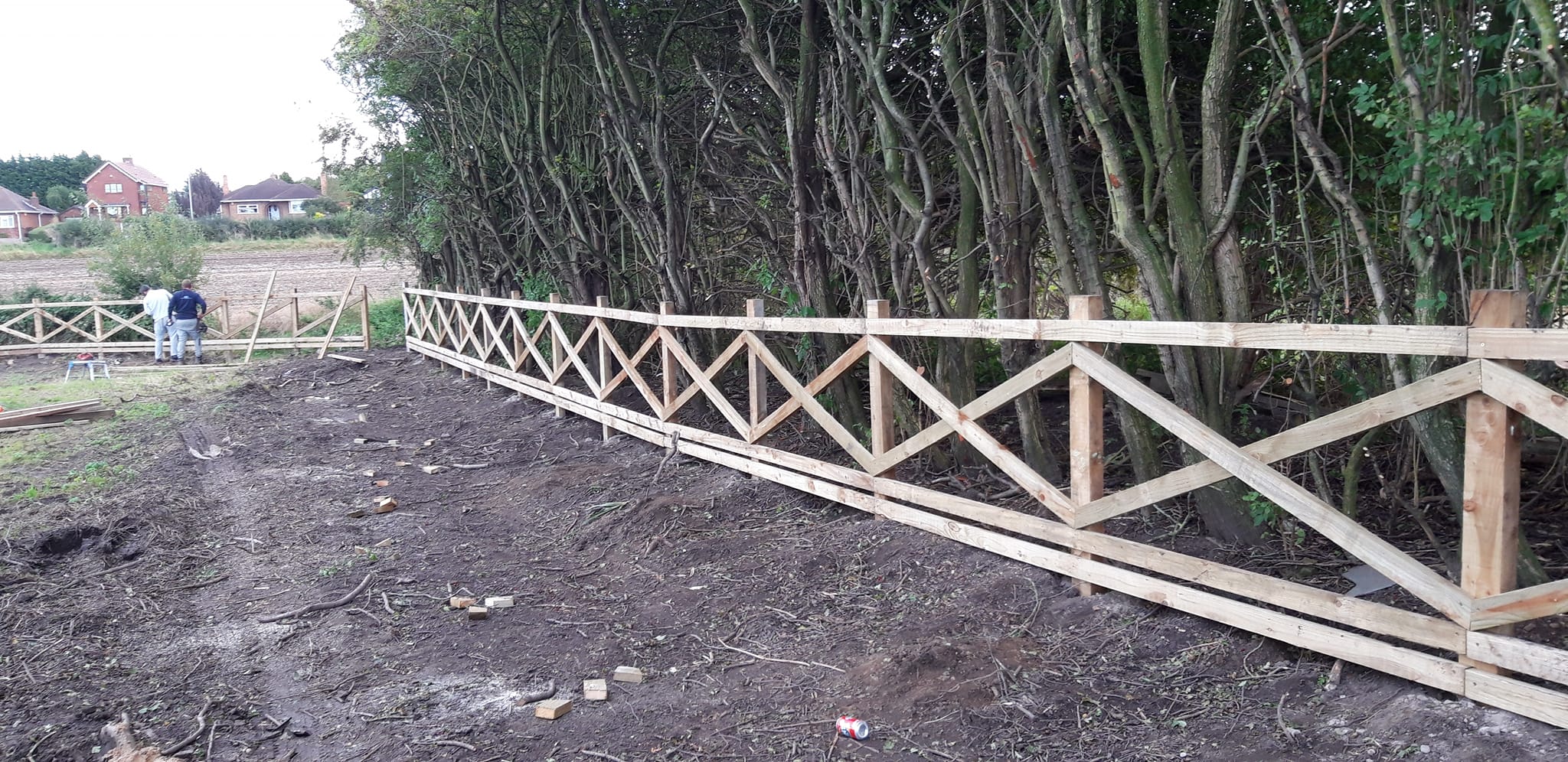 Post and rail fencing installation in rural setting, with two workers assembling wooden panels along a boundary, surrounded by trees and open land.