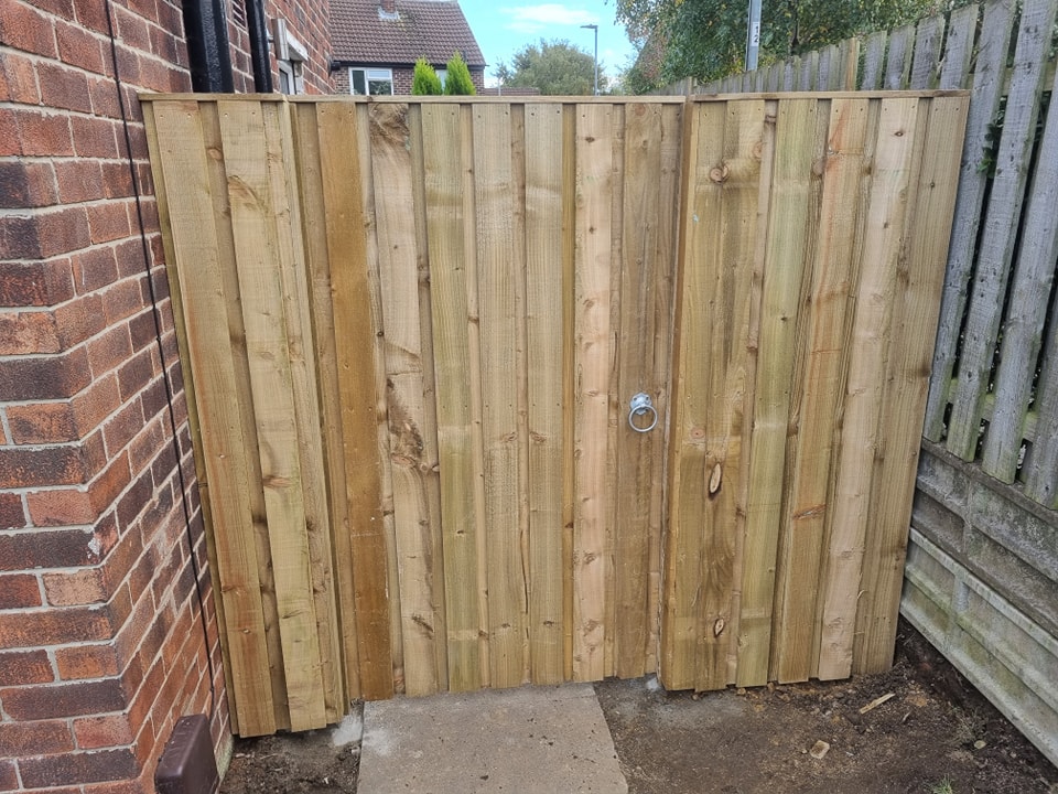 Wooden garden gate installed beside a brick wall, featuring vertical planks, a circular metal handle, and a natural wood finish, emphasizing privacy and security for residential properties.