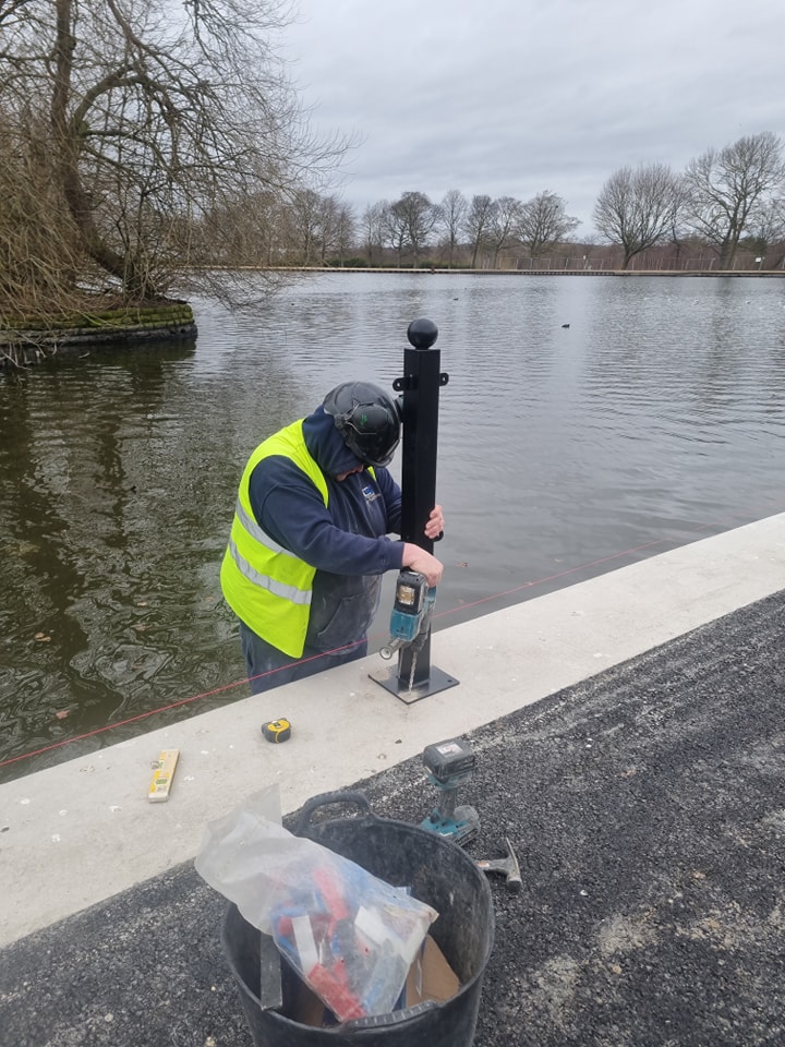 Worker in high-visibility vest installing a fence post near a body of water, with tools and materials visible on the ground, illustrating CK Fencing Contractors' installation services.