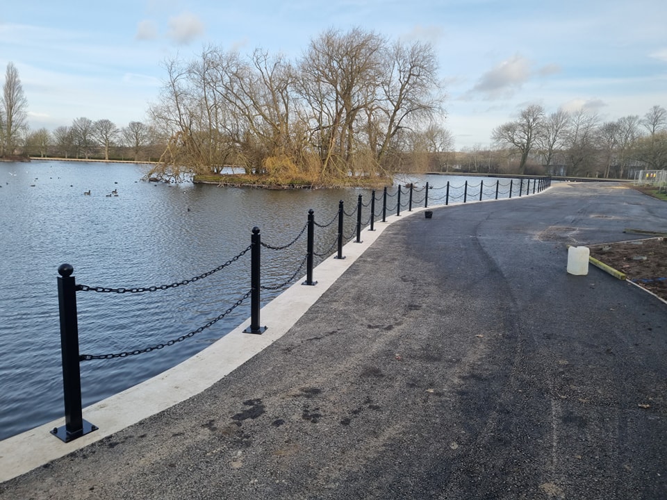 Black chain-link fencing along a water edge, enhancing security and aesthetics for a park area, with trees in the background and ducks swimming in the water.