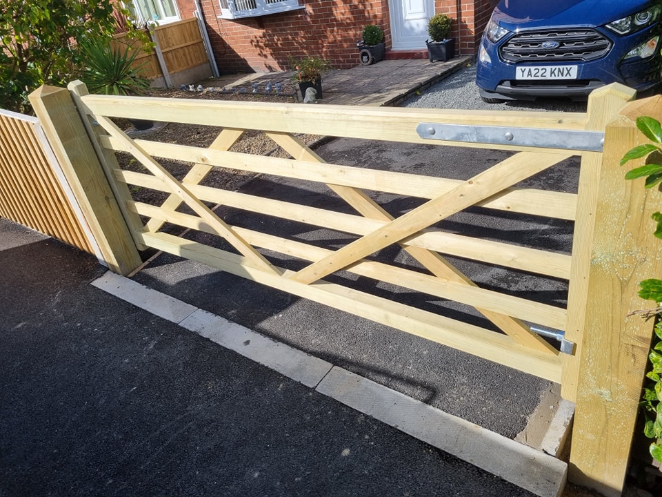Bespoke wooden driveway gate installation at a residential property, showcasing a sturdy design with diagonal slats and metal latch, surrounded by landscaped garden and parked vehicle.