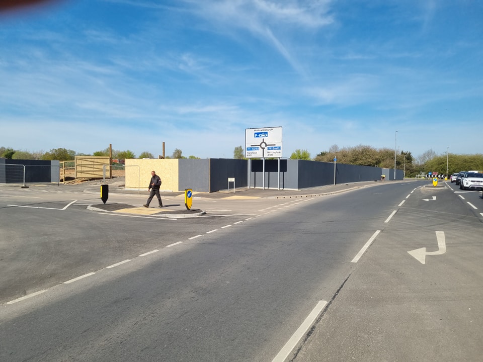 Construction site with temporary hoarding and signage, pedestrian crossing, and a person walking along the road, illustrating urban development and safety measures in outdoor spaces.