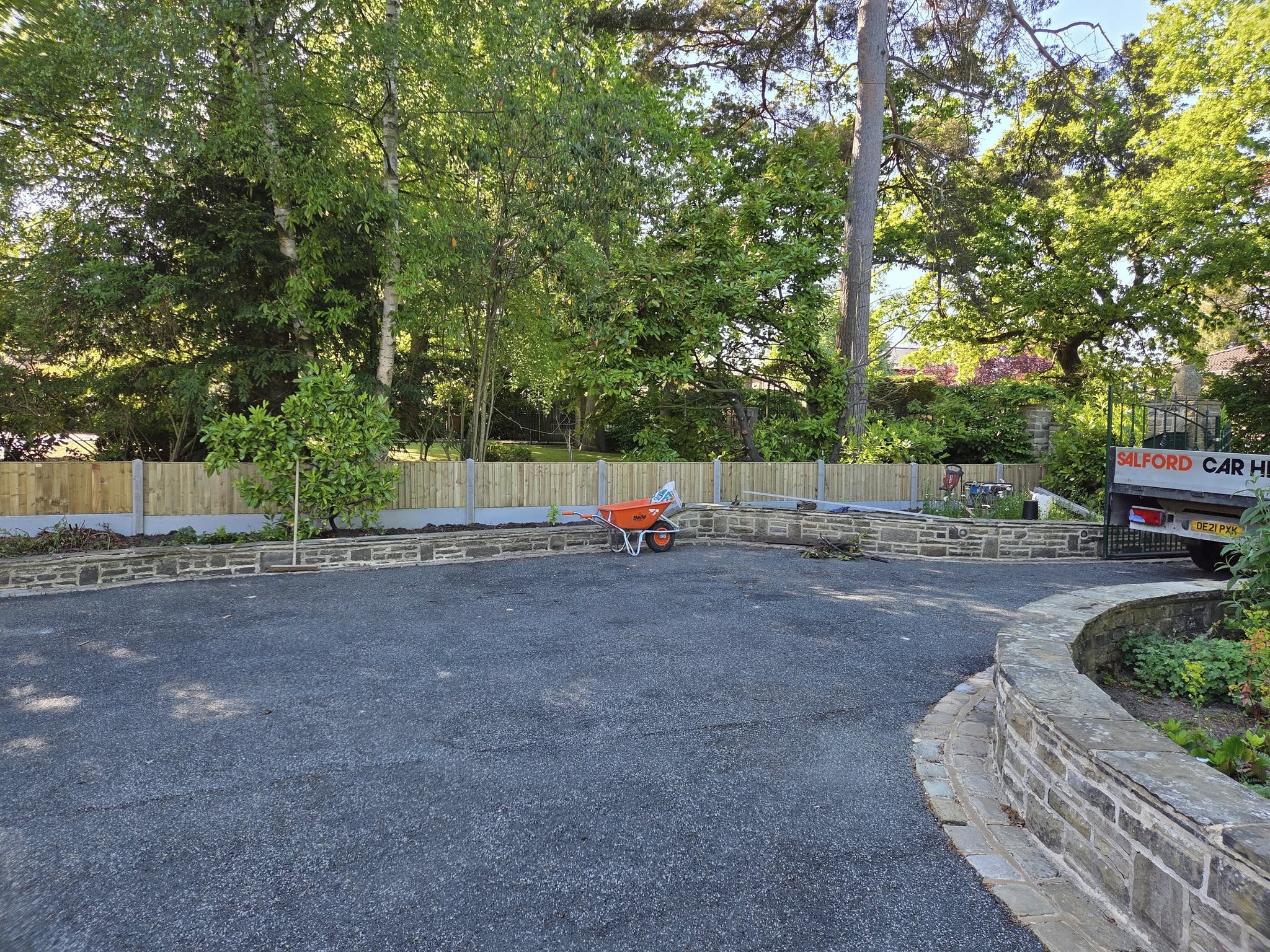 Residential driveway with a wheelbarrow, surrounded by greenery and a stone wall, showcasing a potential area for decking installation by CK Fencing Contractors.