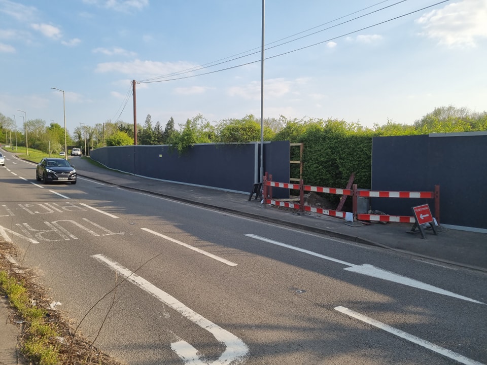 Temporary security hoarding along a roadside, featuring solid blue panels and construction barriers, with a car passing by and greenery in the background.