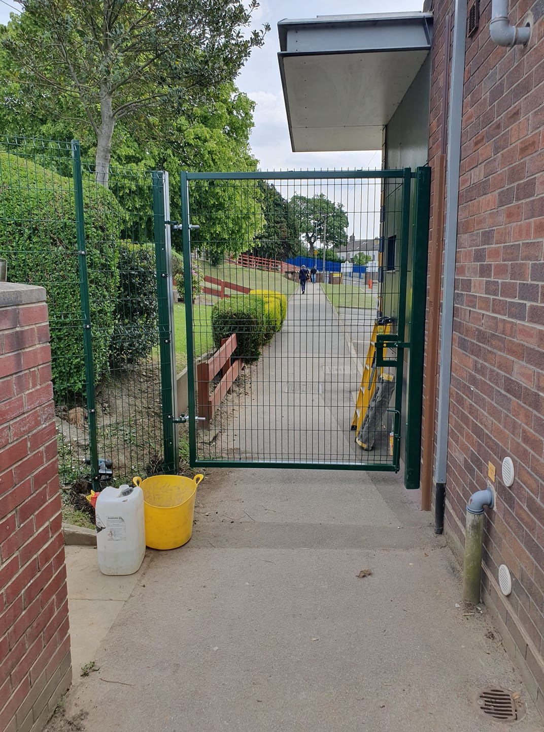Gate entrance to a sports facility with green MUGA fencing, surrounded by greenery, featuring a yellow bucket and a white container on the ground, leading to a pathway.