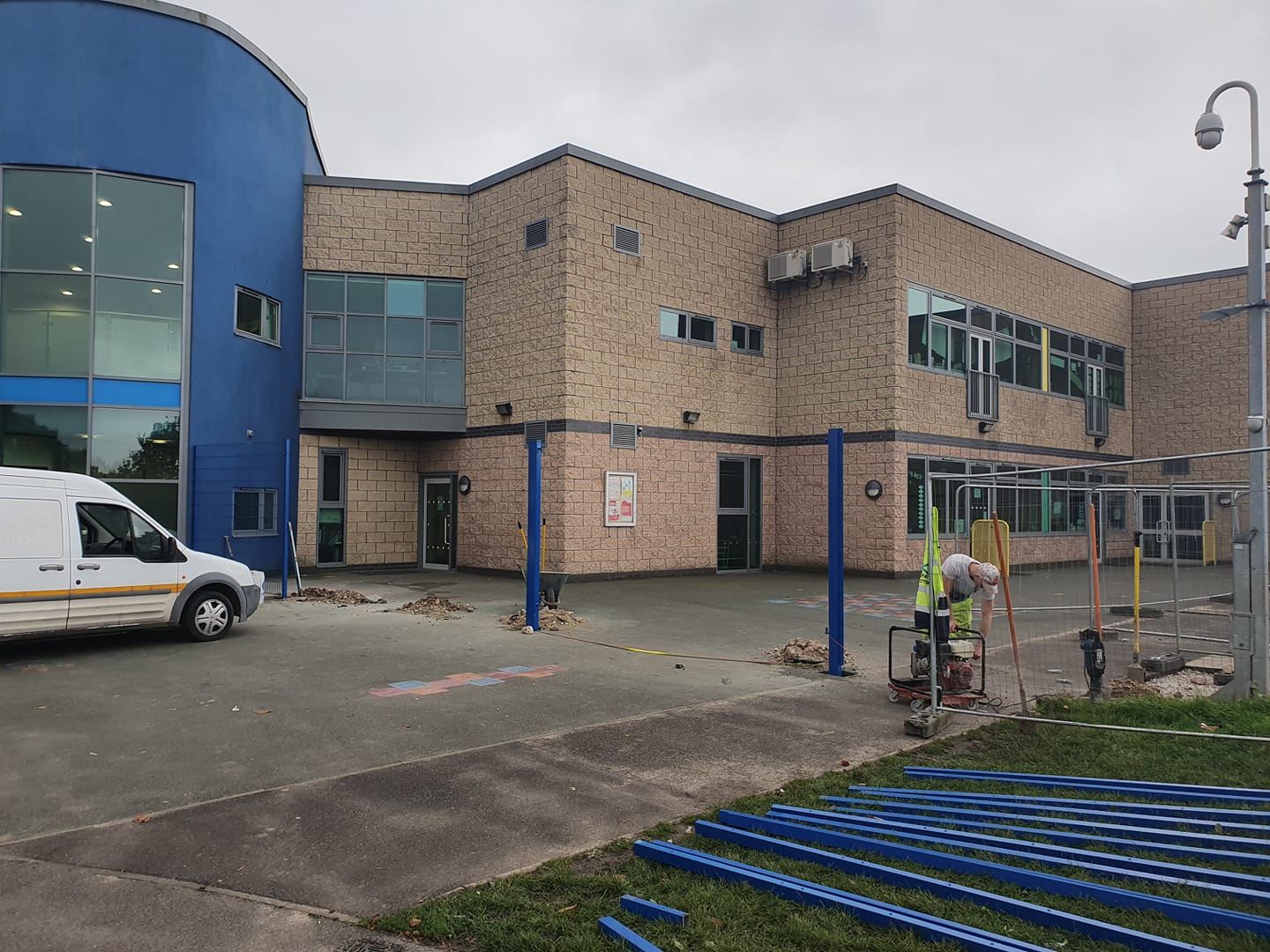 Construction site of a sports pitch with a worker operating machinery, blue fencing materials, and a van, near a modern building, reflecting CK Fencing Contractors' installation services for MUGA and sports pitch fencing.