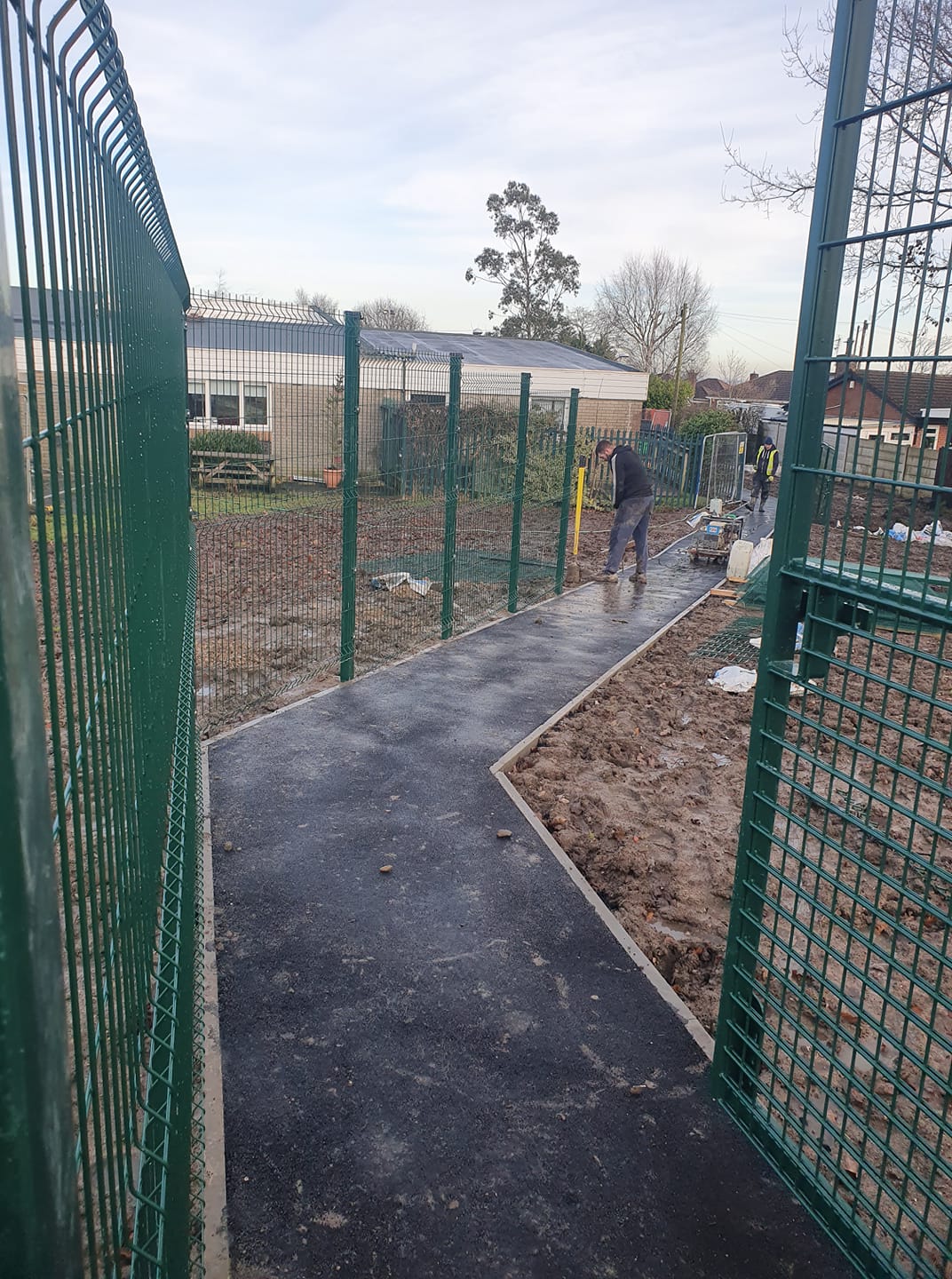Installation of green playground fencing along a newly paved pathway in a play area, featuring construction workers and safety measures, emphasizing compliance with UK safety standards.