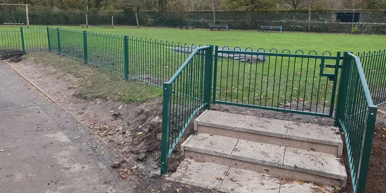 Playground fencing with green metal railings enclosing a grassy area, featuring steps leading into the play space, emphasizing safety and compliance with UK standards.