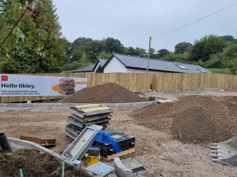 Construction site featuring fencing installation for McDonald's in Ilkley, with a wooden fence, piles of gravel, and construction materials visible.