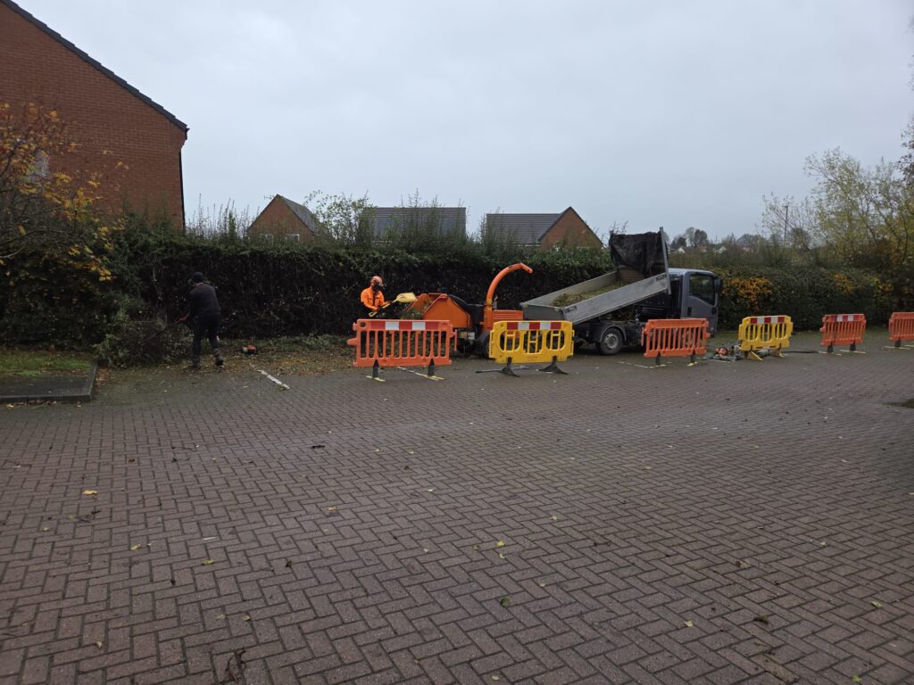 Workers using machinery for hedge trimming and landscaping maintenance, with orange barriers in a residential area.