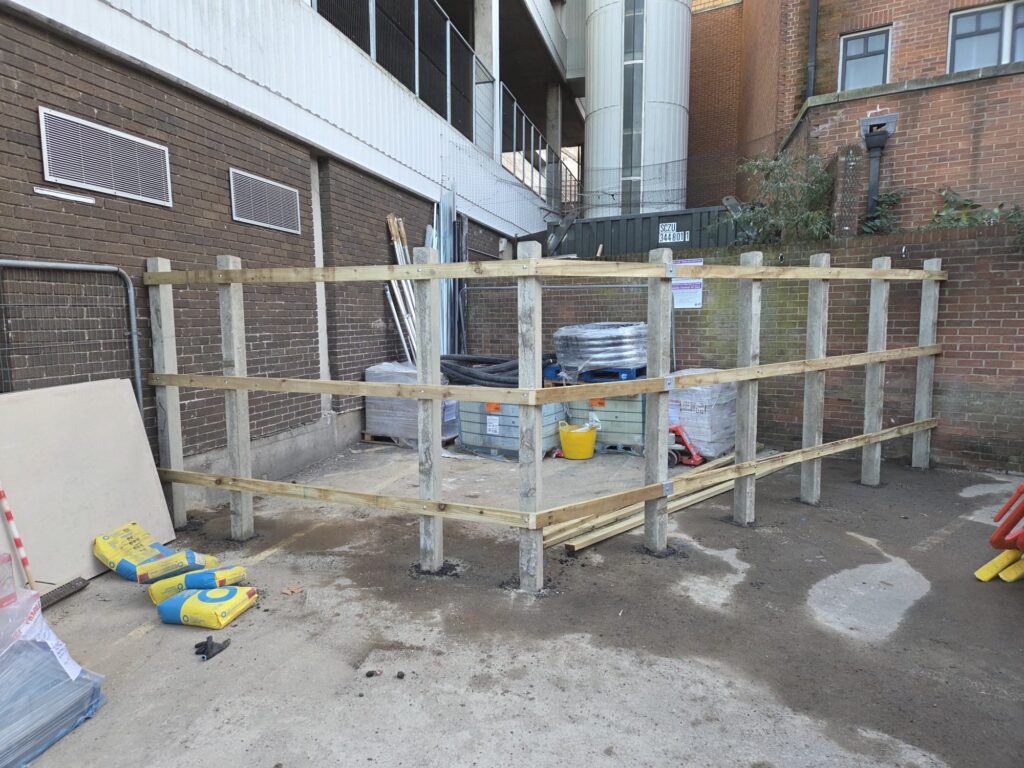 Site hoarding installation at CFA London, featuring wooden posts and horizontal beams, with construction materials and tools visible in the background.