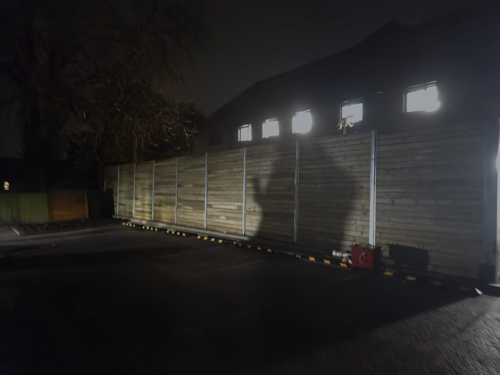 Wooden site hoarding illuminated at night, showcasing shadows and structural details, relevant to CK Fencing Contractors' fencing solutions for commercial projects.