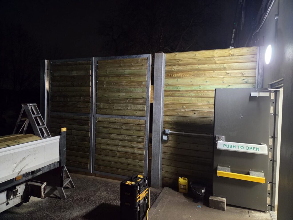 Heavy-duty acoustic fencing installation with a metal frame and wooden panels, featuring a service door labelled "PUSH TO OPEN," adjacent to a work vehicle and a ladder, showcasing CK Fencing Contractors' expertise in secure fencing solutions.